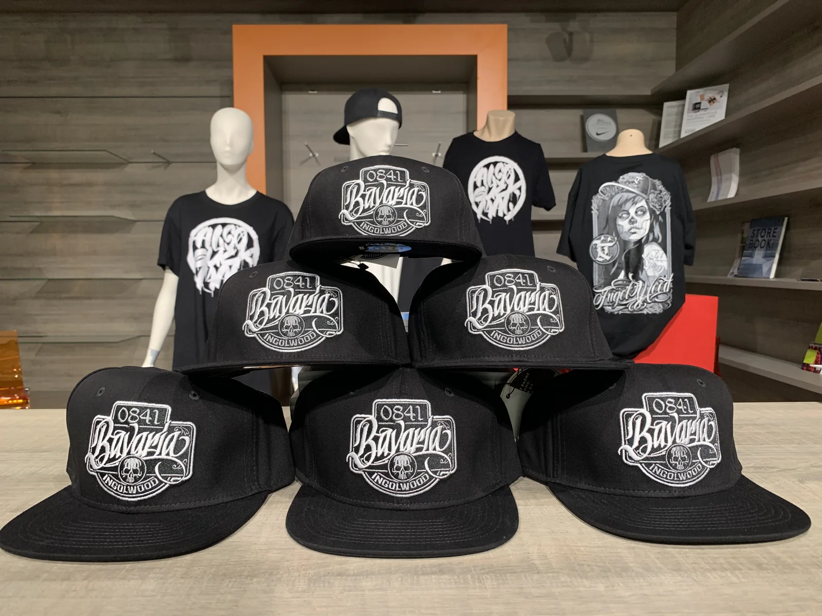 Gestapelte Snapback-Caps mit Bavaria-Patch auf einem Tisch, dahinter Shirts auf Schaufensterpuppen