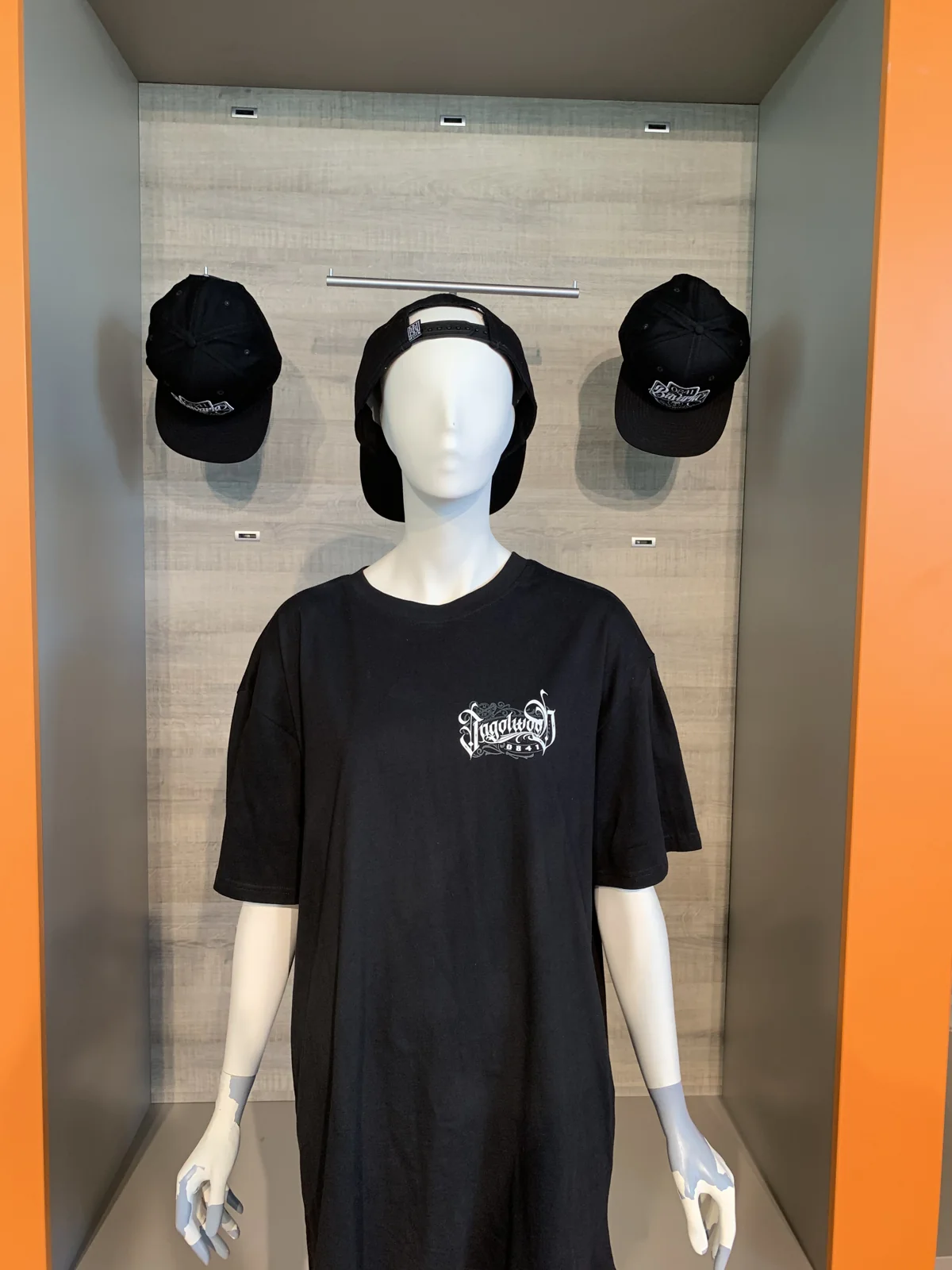 Schwarzes T-Shirt mit kleinem Ingolwood-Logo auf der Brust, Caps an der Wand