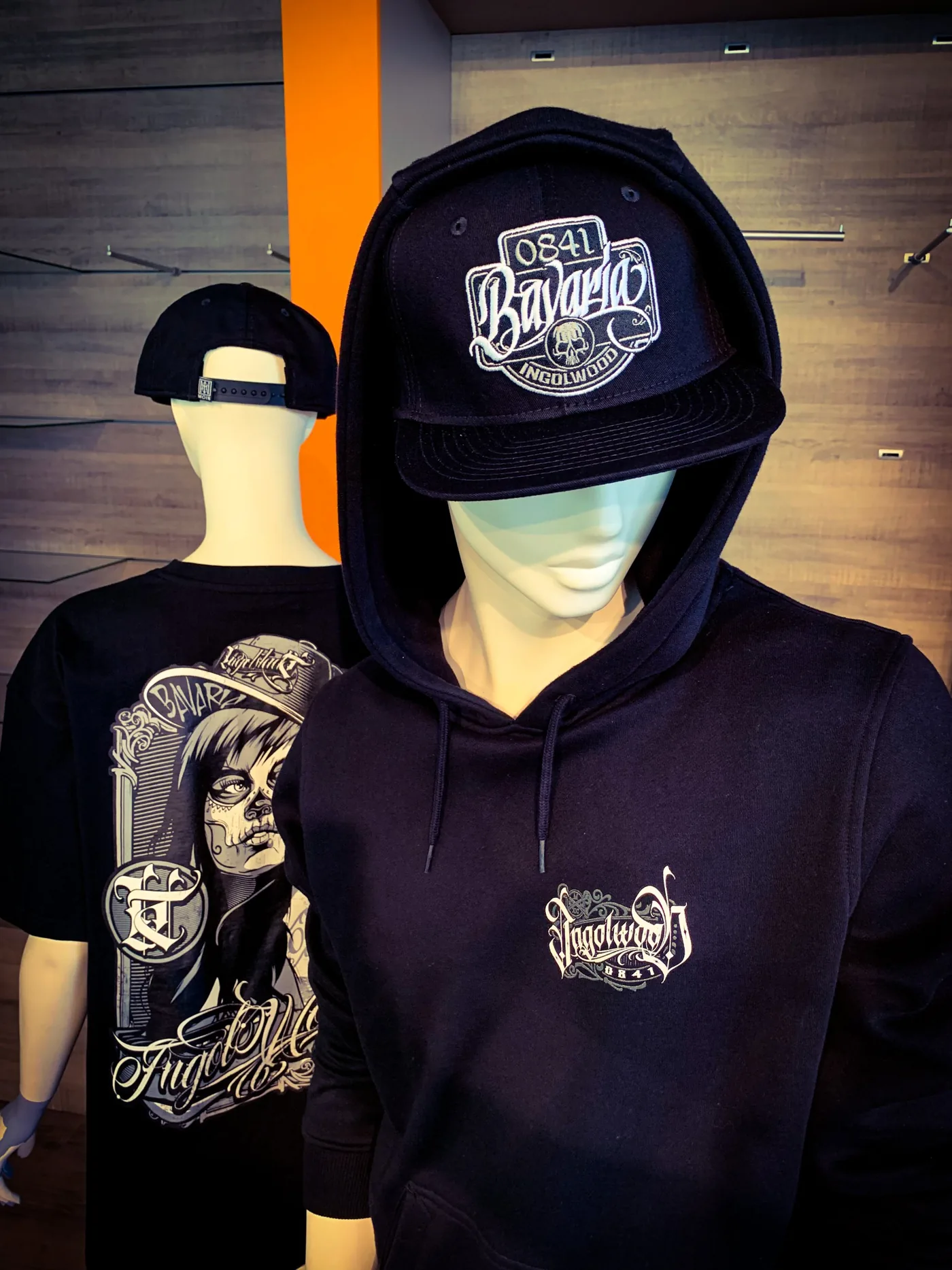 Schwarzer Hoodie und Snapback-Cap mit Bavaria-Patch auf Schaufensterpuppen, daneben ein Shirt mit Rückenmotiv